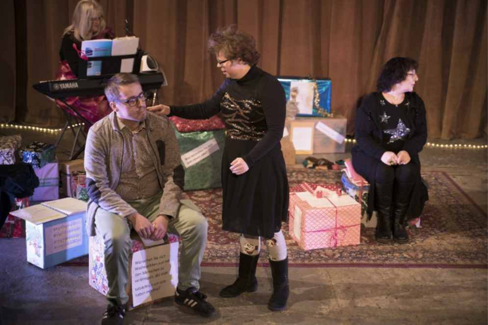 Das Foto zeigt eine Theaterszene: Vier Menschen sitzen oder stehen auf einer Bühne.